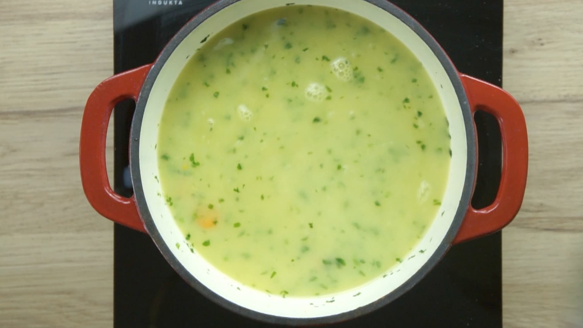 Carrot slices, parsnip slices, and chopped green herbs are in a creamy, yellow soup in a red enameled Dutch oven.