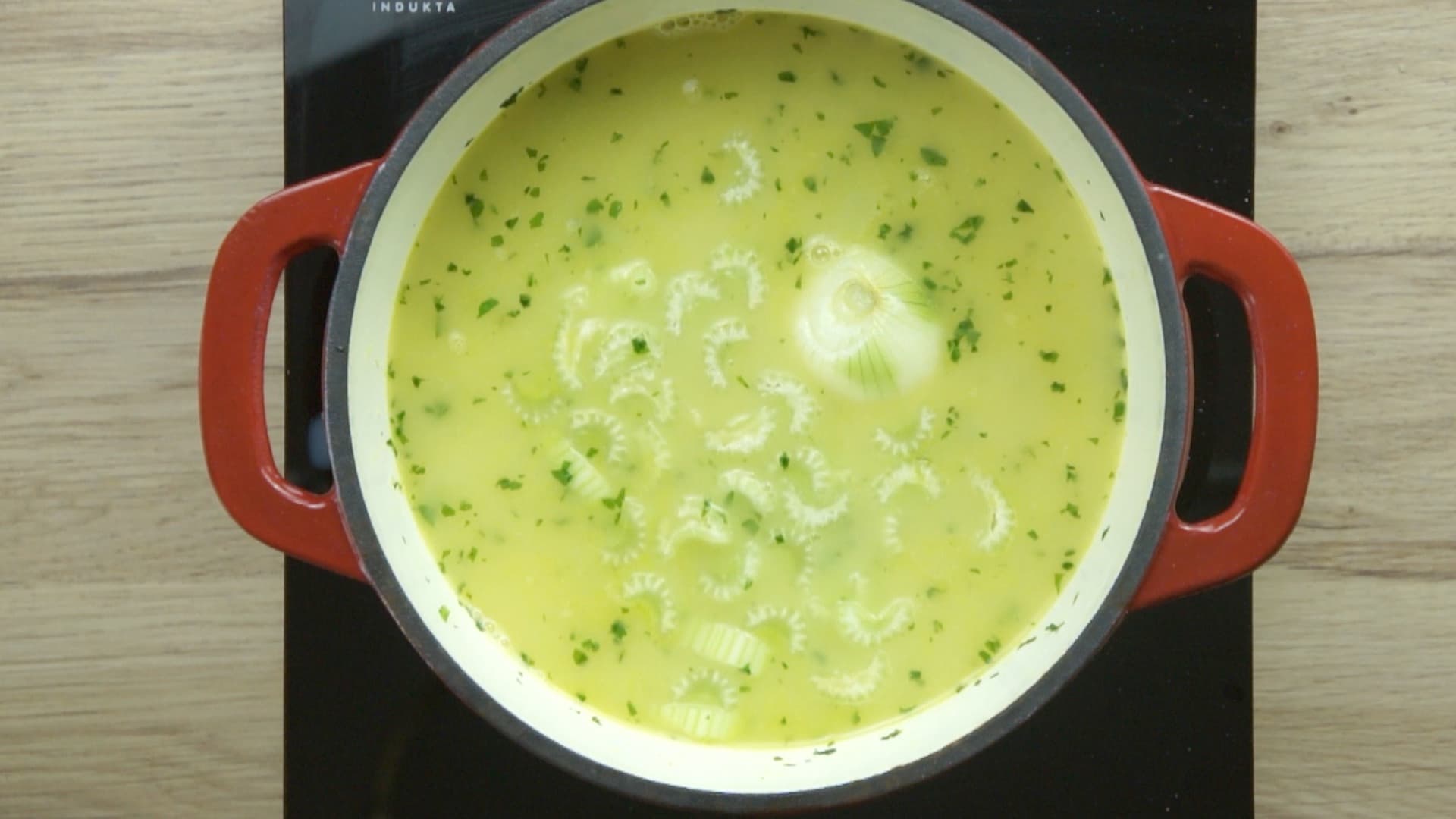 Carrot slices, parsnip slices, whole onion, and chopped celery stalk, and freshly chopped green herbs are in a light yellow soup in a red enameled Dutch oven.