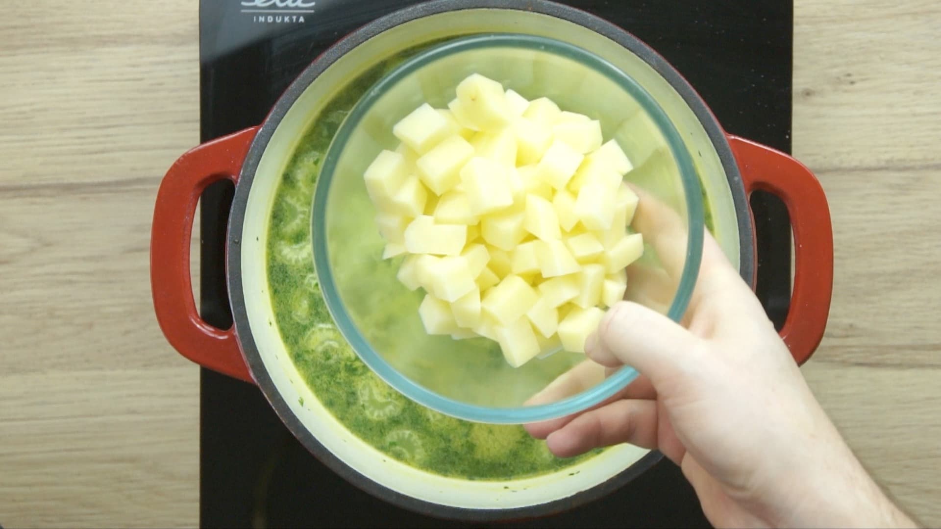 A hand is holding a bowl of chopped potatoes to be added to a light yellow soup below.