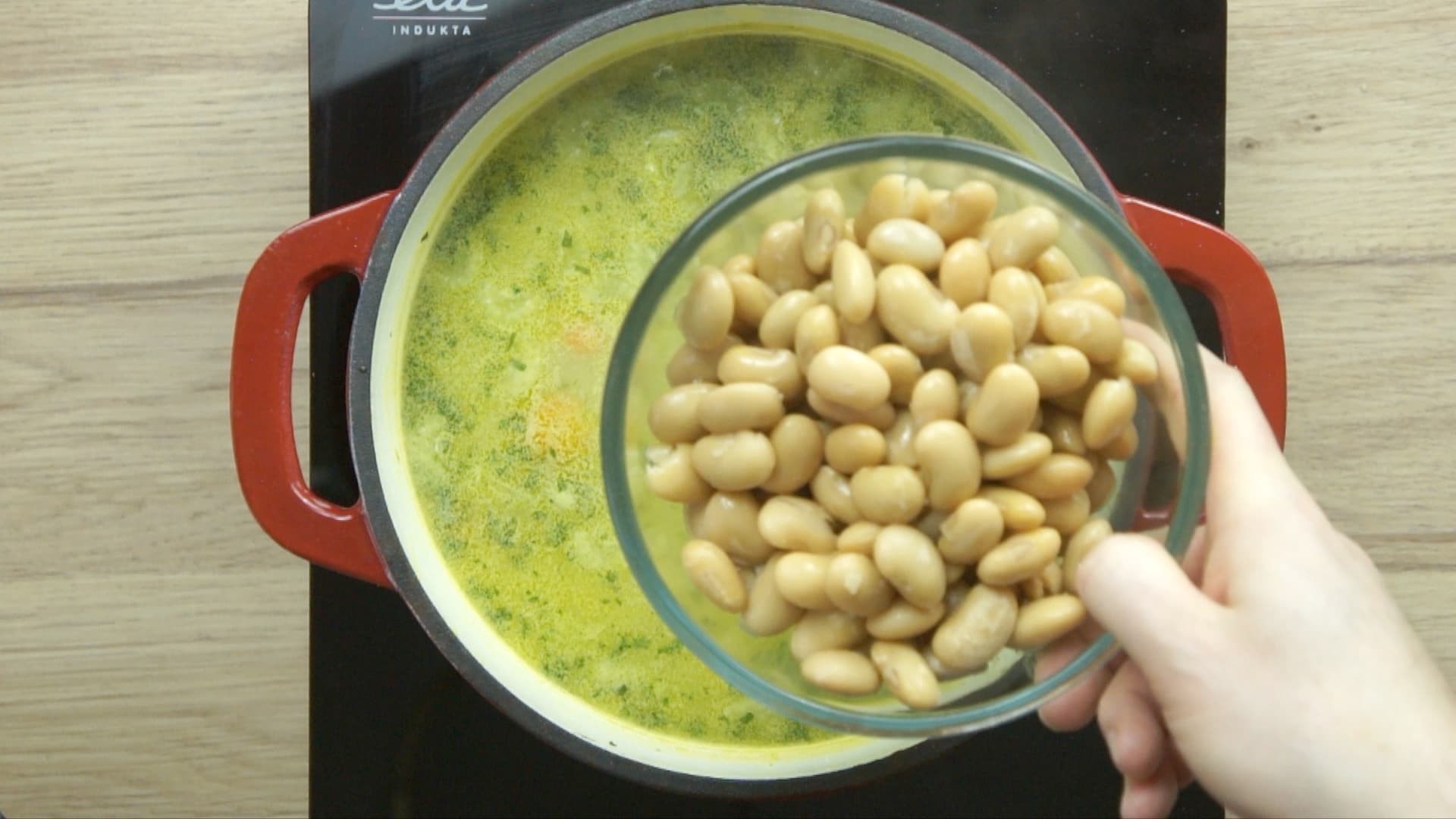 A hand is holding a bowl of chopped white beans to be added to a light yellow soup below.