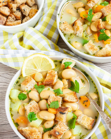 Light yellow soup served in two white bowls full of large white beans, croutons, carrot slices, and diced potatoes. It is sprinkled with freshly chopped herbs.