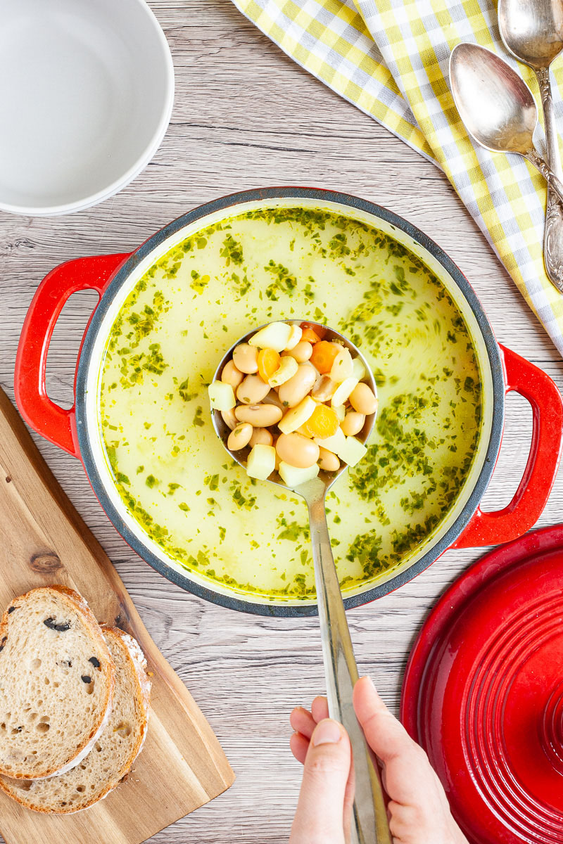 Yellow soup with chopped herbs is served in a large red dutch oven. A laddle is full of white beans, diced potatoes and carrot slices as it takes one serving.