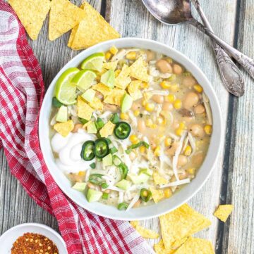 A white bowl with a light brown chili with white beans, shredded mushrooms, corn, topped with green chili slices, shredded cheese and nacho chip pieces.