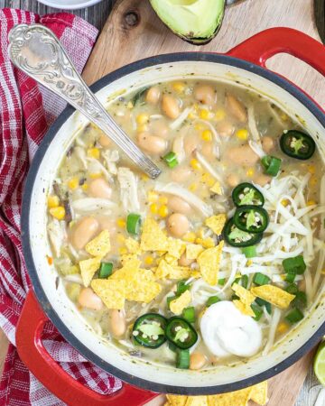 A red enameled Dutch oven with a light brown chili with white beans, shredded mushrooms, corn, topped with green chili slices, shredded cheese and nacho chip pieces. Avocado, and other toppings are around it.