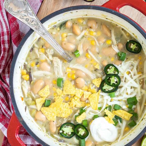 A red enameled Dutch oven with a light brown chili with white beans, shredded mushrooms, corn, topped with green chili slices, shredded cheese and nacho chip pieces. Avocado, and other toppings are around it.