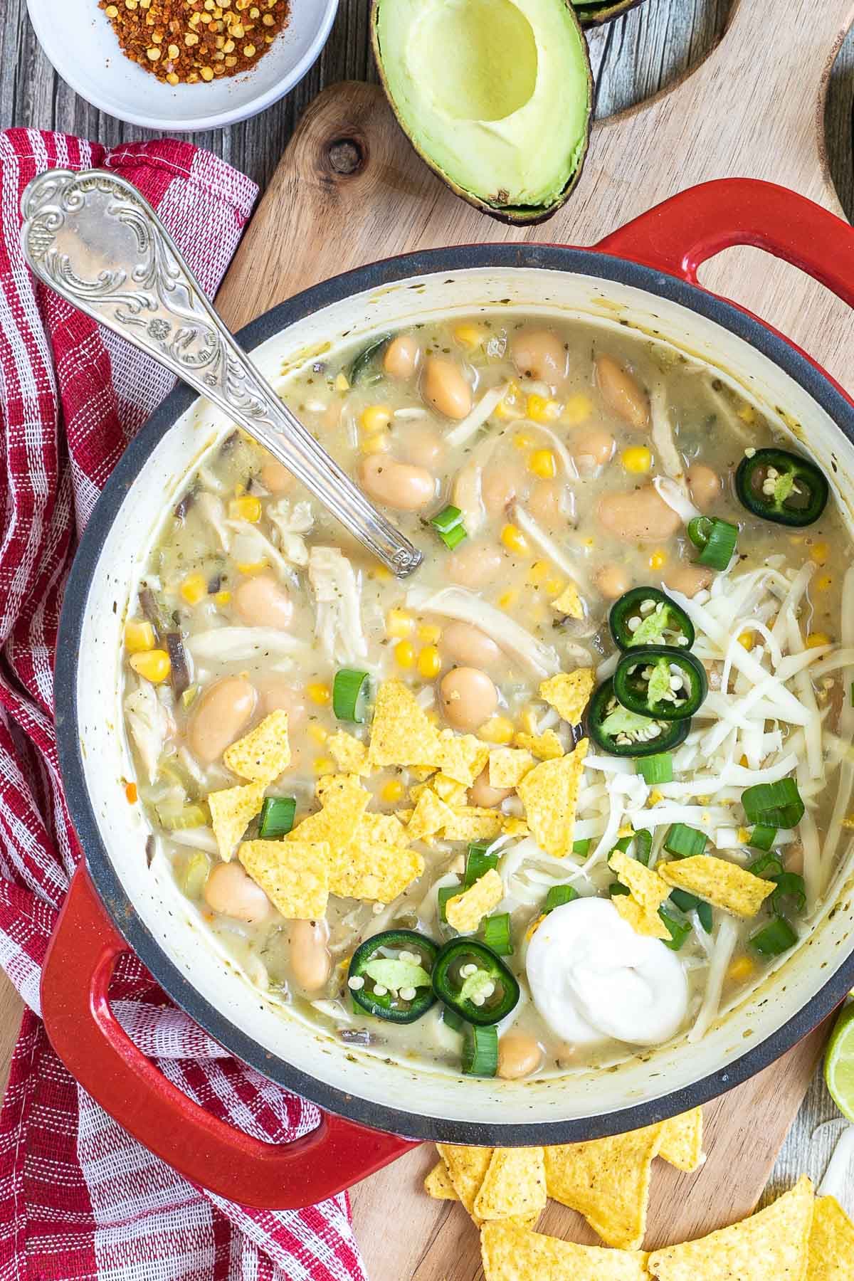 A red enameled Dutch oven with a light brown chili with white beans, shredded mushrooms, corn, topped with green chili slices, shredded cheese and nacho chip pieces. Avocado, and other toppings are around it.