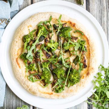 A pizza on a white pizza plate topped with white sauce, zucchini, broccoli, asparagus, mushrooms, and fresh arugula.