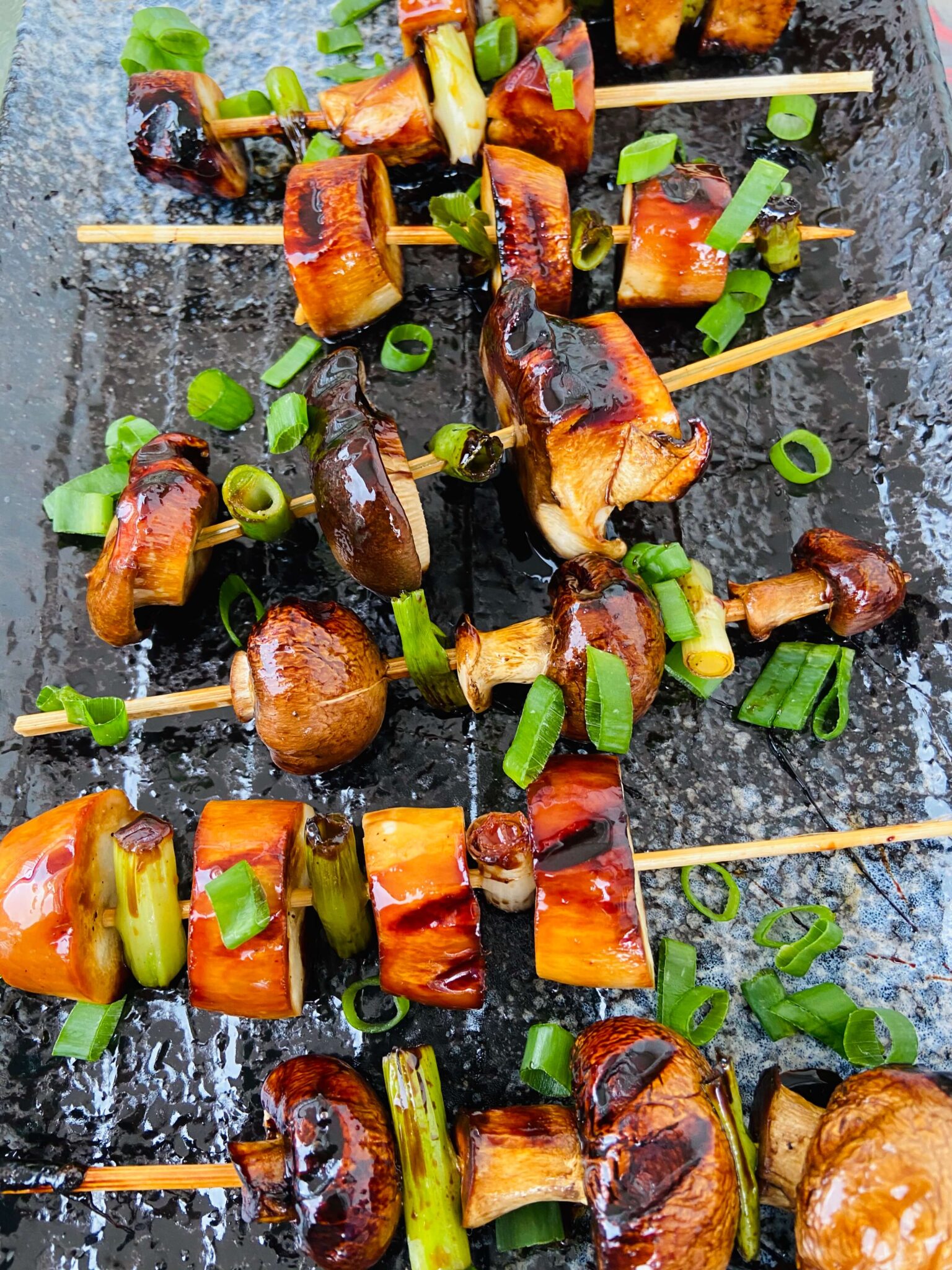 Wooden skewers with mushroom slices grilled to dark brown almost black on the edges. They are sprinkled with green onion rings