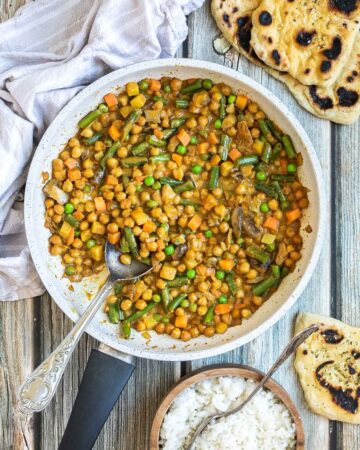 White frying pan full of vegetables like chickpeas, green beans, chopped carrots, green peas, chopped mushroom, and potatoes swimming in a brown-orange sauce. Naan bread and a brown bowl of rice are next to it.