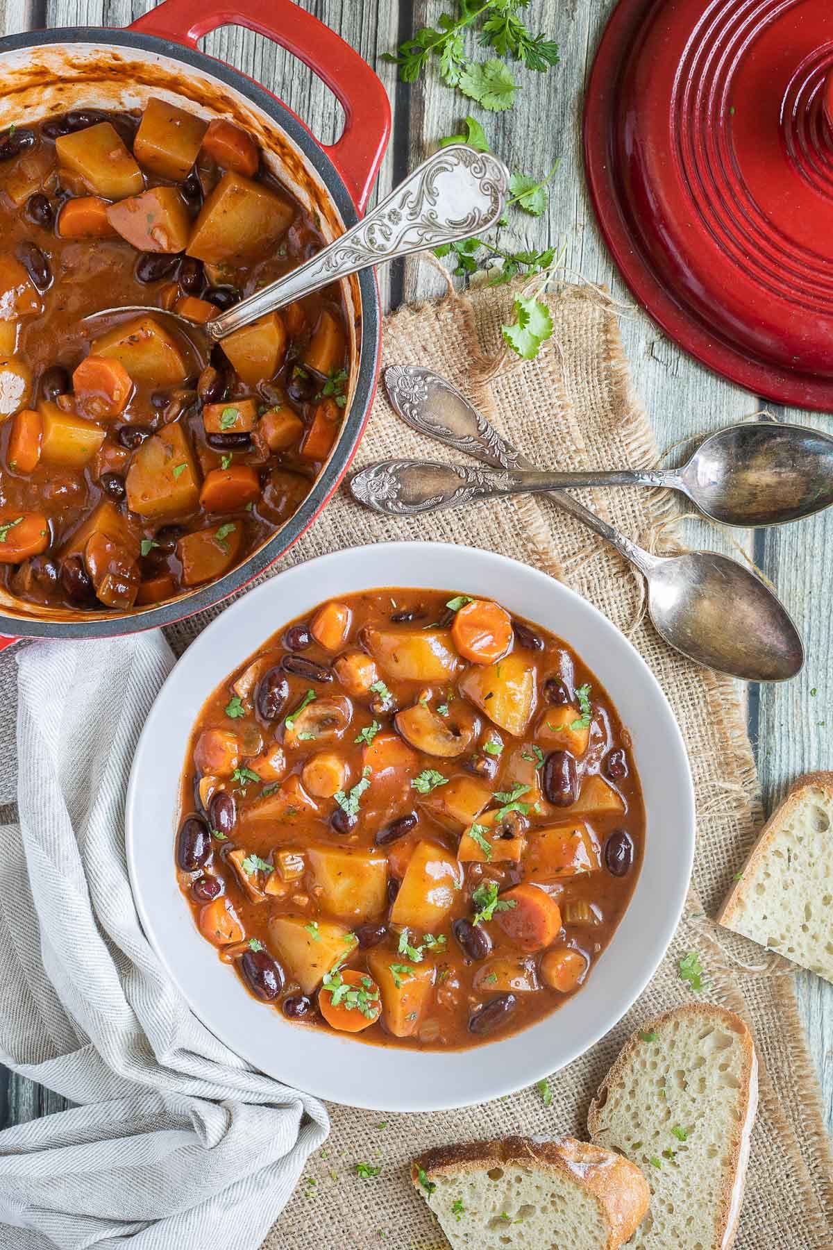 Vegetable stew in a white bowl with lots of chopped vegetables like potatoes, carrots, mushroom, celery and red kidney beans. Topped with freshly chopped green herbs. Leftover stew is next to it as well as two spoons.