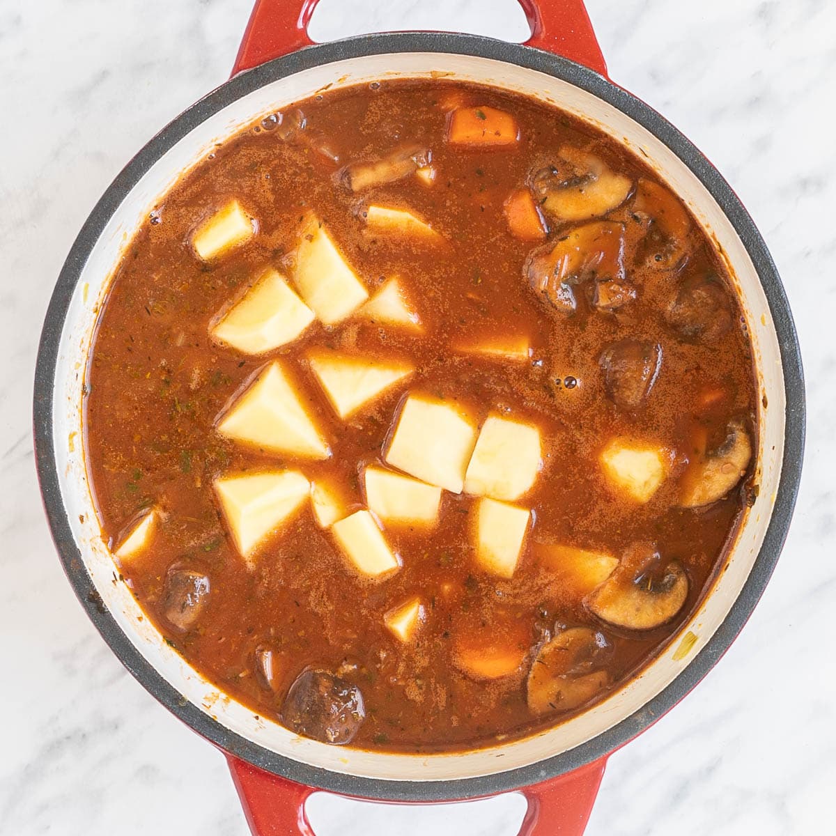 Red white enameled Dutch oven with a red thick sauce, lots of chopped veggies like carrots, mushrooms, celery and raw potatoes in the middle.