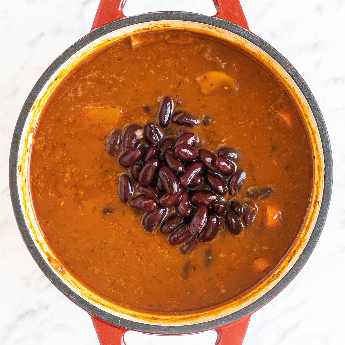 Red white enameled Dutch oven with a red thick sauce, lots of chopped veggies like carrots, mushrooms, celery and red kidney beans in the middle.