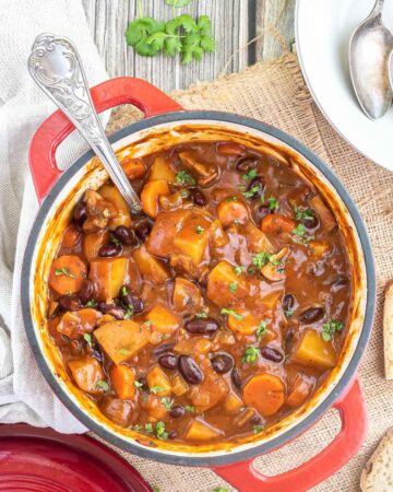 Vegetable stew in a red white enameled Dutch oven with lots of chopped vegetables like potatoes, carrots, mushroom, celery and red kidney beans. Topped with freshly chopped green herbs.