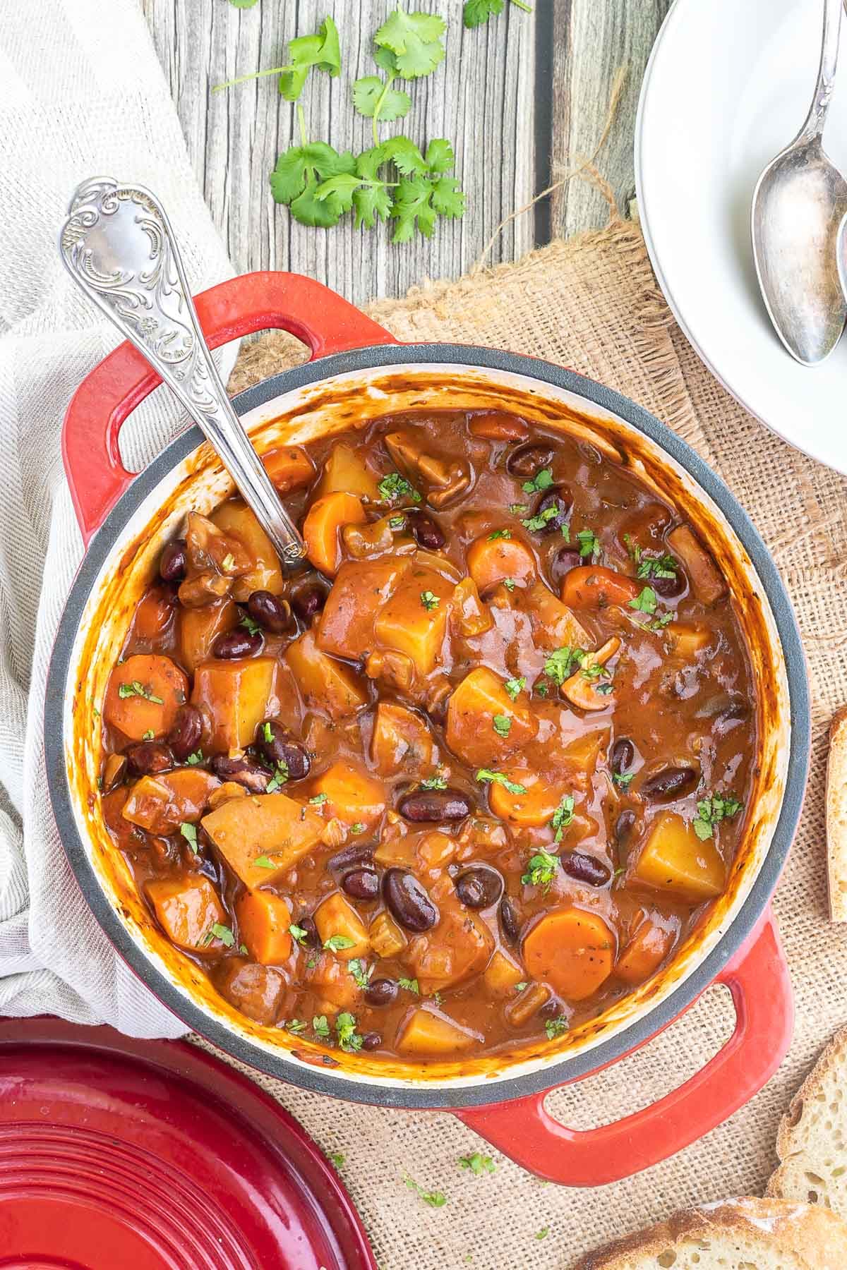 Vegetable stew in a red white enameled Dutch oven with lots of chopped vegetables like potatoes, carrots, mushroom, celery and red kidney beans. Topped with freshly chopped green herbs.