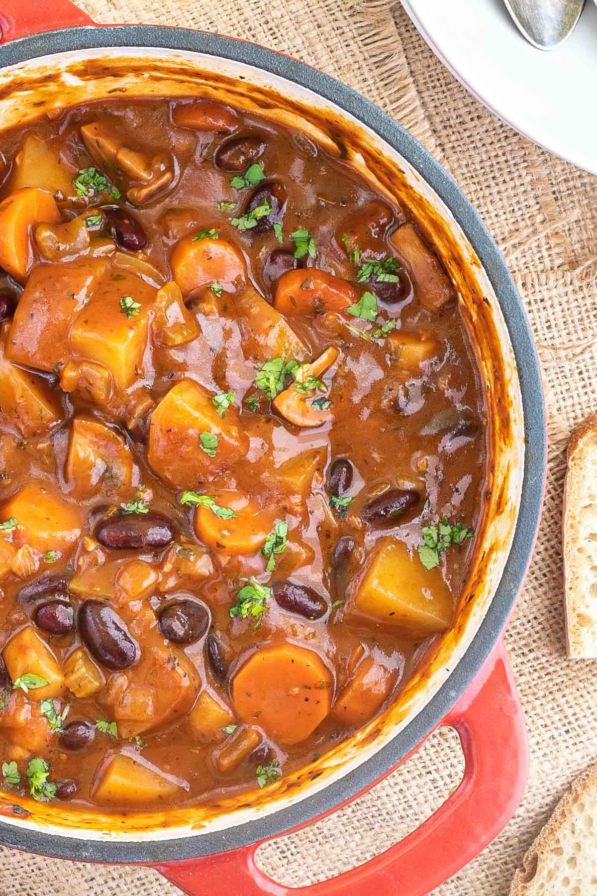 Vegetable stew in a red white enameled Dutch oven with lots of chopped vegetables like potatoes, carrots, mushroom, celery and red kidney beans. Topped with freshly chopped green herbs.