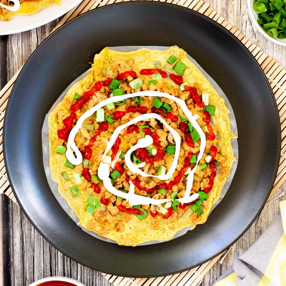 Black plate with opaque rice paper topped with a yellow batter and sprinkled with brown crumbles and green spring onion rings. White and red sauces are drizzled on top.