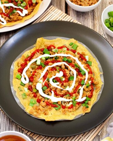 Black plate with opaque rice paper topped with a yellow batter and sprinkled with brown crumbles and green spring onion rings. White and red sauces are drizzled on top.