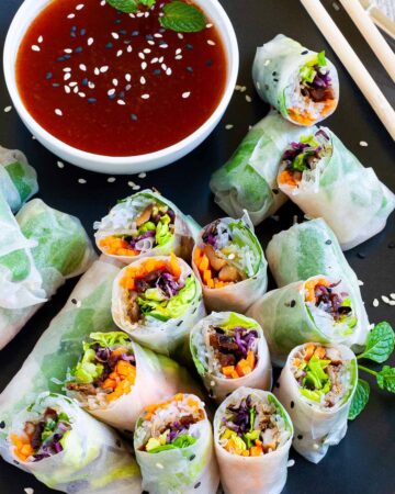 Opaque rice paper rolls filled with carrots, purple sprouts, thin glass. noodles and brown mushrooms are served on a large black plate. Several rolls are cut half so you can see the filling. Red sauce in a small white bowl is served next to it.