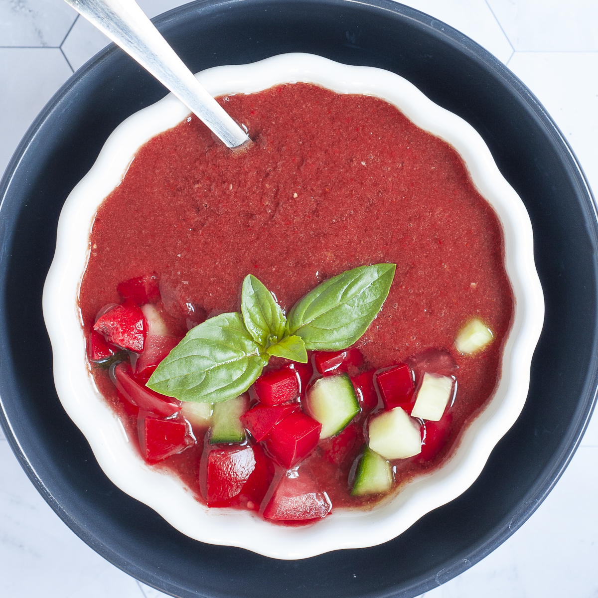 Red soup in two white and black bowls topped with basil leaves and diced red and green vegetables.