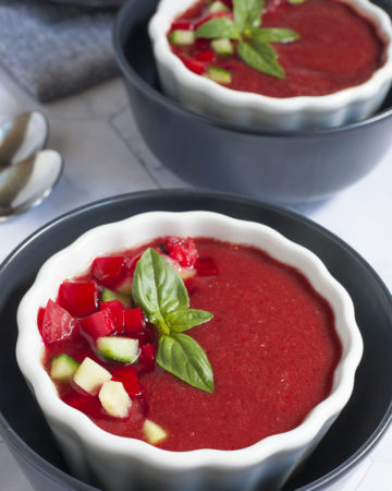 Red soup in 2 white and black bowls topped with basil leaves and diced red and green veggies.