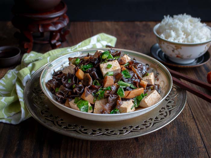 Silver plate with a white bowl filled with tofu cubes, chopped green leaves, carrot slices and brown wood ear mushroom pieces.