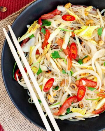 Black plate on a red mate with thin glass noodles, sliced tomatoes, roasted peanuts, sliced onion, sliced chili peppers, chopped green herbs. Chopsticks are placed on the side.