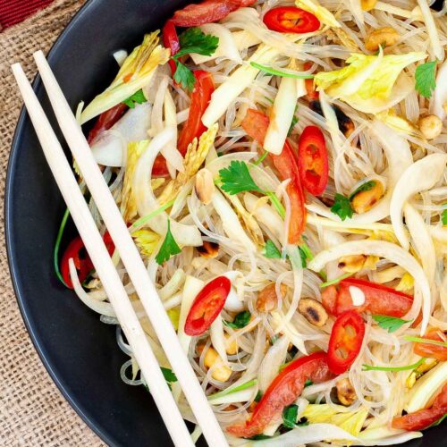 Black plate on a red mate with thin glass noodles, sliced tomatoes, roasted peanuts, sliced onion, sliced chili peppers, chopped green herbs. Chopsticks are placed on the side.