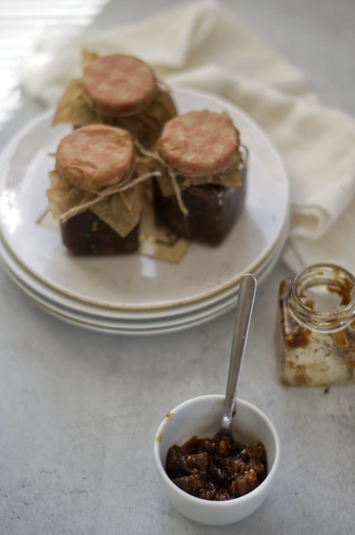 A bowl of chutney surrounded by an empty glass jar and three full jars on a table with a white cloth.