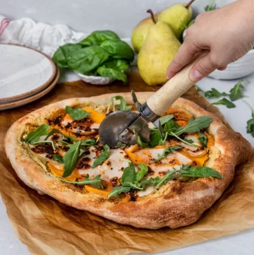 Pizza with butternut squash slices, pear slices and fresh leafy greens. A hand is holding a pizza cutter to cut into slices.